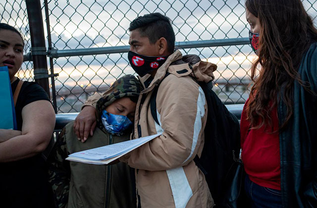 Immigration. Latinix father wearing tan coat and black face mask stands in front of gate with young boy leaning on him, also wearing a mask, and Latinx woman wearing red shirt and black jacket, with a face mask.