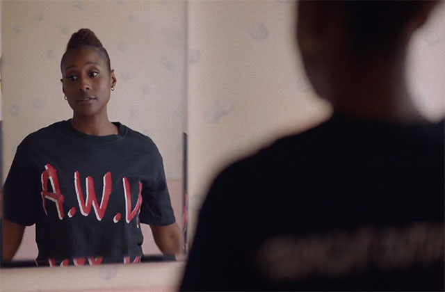 Insecure. Black woman looking in mirror at herself in bathroom, wearing black N.W.A. tee shirt.