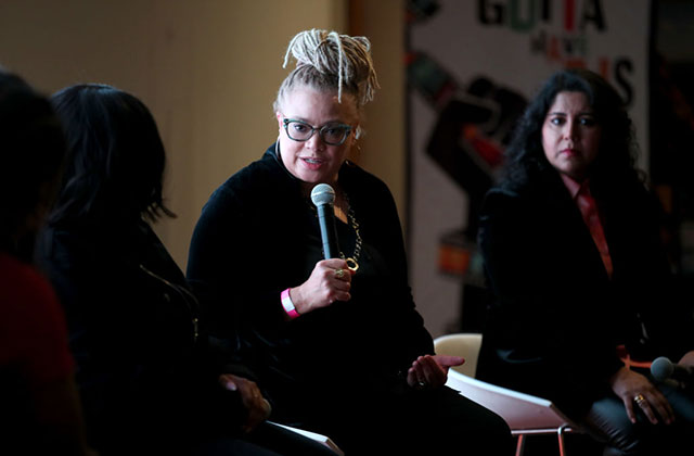 Kasi Lemmons. Black woman with blonde locs in top bun seated, wearing all black holding a microphone.