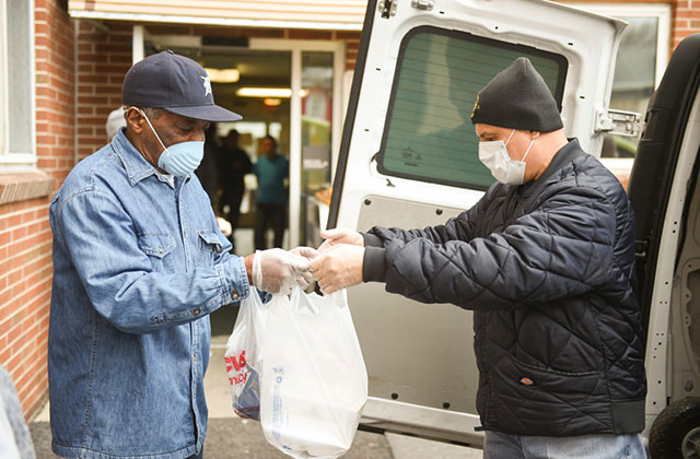Latinx Community. Elderly man wearing blue shirt and baseball cap accepts food from the back of a white truck, where a man in black stands handing out food.