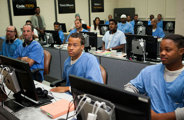 A group of Black men wearing blue prison uniforms sit in front of computers in a classroom setting.