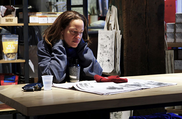 Drew Dixon. Black woman with shoulder length brown hair wearing blue sweatshirt sitting alone at table in coffee shop.