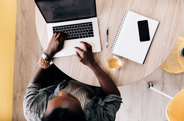 Positive Peers. Aerial shot of Black man working at a laptop with smartphone and white notepad on table, next to golden colored drink.