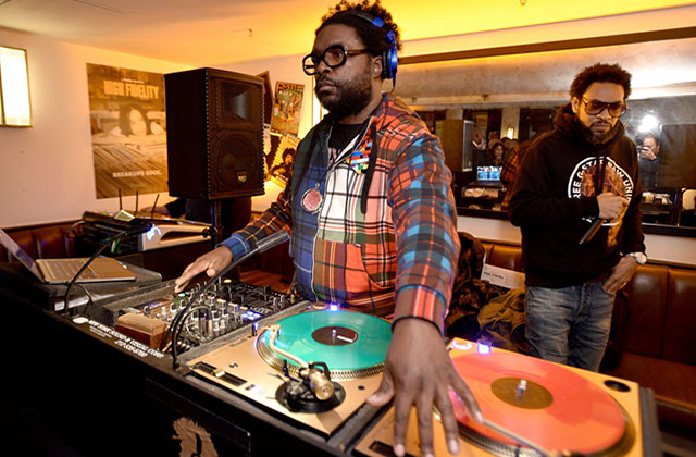 Questlove. Black man with short dark hair DJ'ing, wearing headphones, an orange and blue plaid shirt and dark framed glasses.