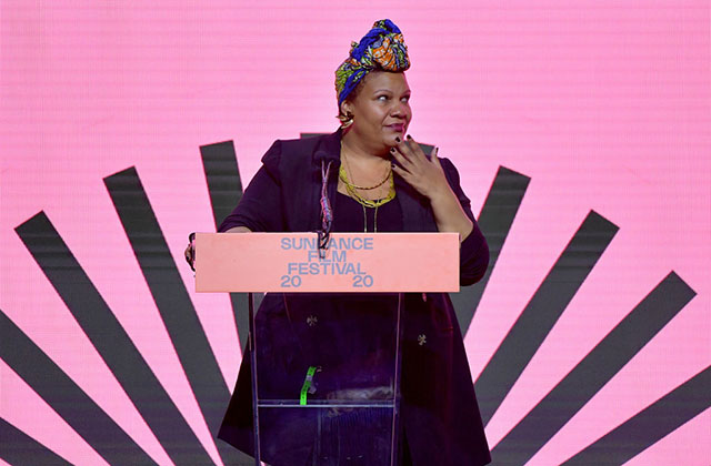 Radha-Blank. Black woman wearing colorful head wrap and dark purple dress, standing in front of podium that reads Sundance Film Festival 2020 and a pink background.