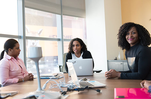 Small Business. Three women of color sit at a conference table with large windows and computers.