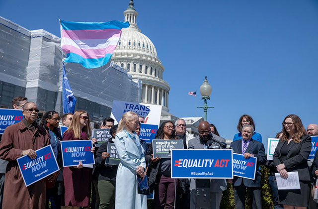 Trans health. A cluster of people in winter coats standing around a podium holding blue signs with the trans flag waving in the background in front of the U.S. Capitol.