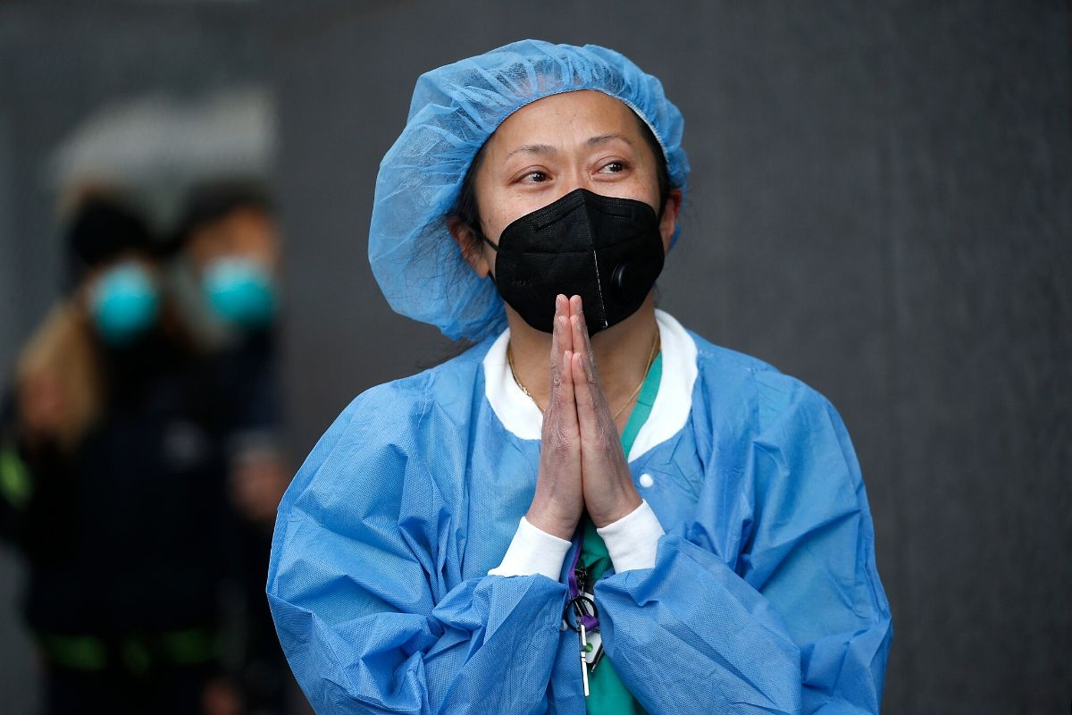 Asian Nurse with blue scrubs and head covering and black mask with hands clasped together in front of her in prayer position. 