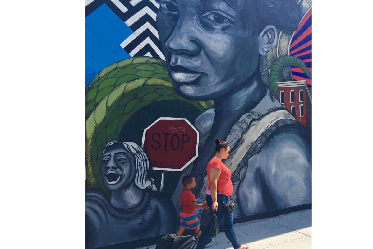 A Latinx mother and child with bright red shirts walk past wall-high mural with Black Latinx woman and child.