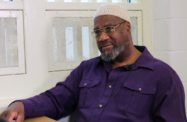 Jalil Muntaqim. Older Black man wearing white kufi and purple shirt and glasses.