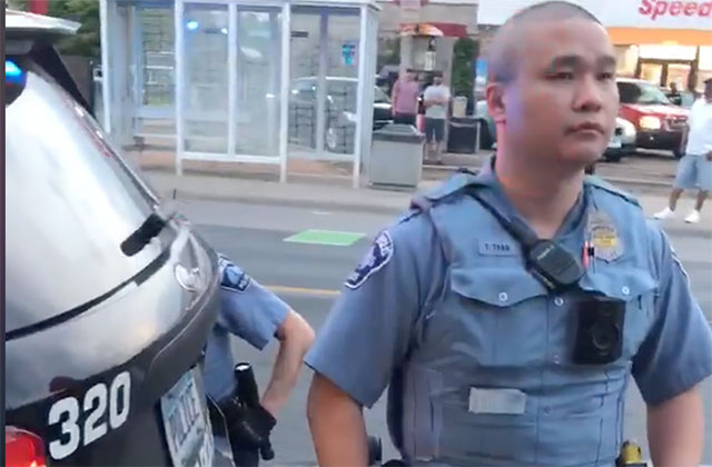 Asian police officerwith a shaven head wearing a blue uniform standing in front of a black police cruiser.