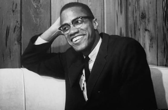 Malcolm X. Archival black and white photo of Black man wearing dark suit, white shirt and black tie.