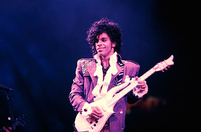 Prince. Black man with long dark hair wearing a purple pant suit, white blouse and playing a guitar on stage.