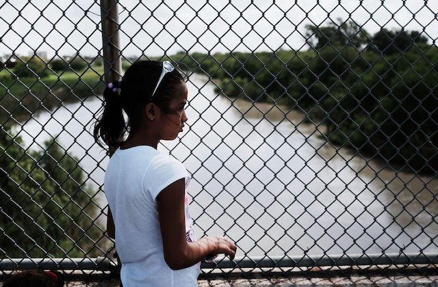 Young girl with dark hair stands in front of a wire fence that overlooks a stream of water.