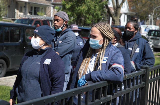 A group of postal workers in uniform gather on the sidewalk to honor a fellow colleague that died of COVID-19.