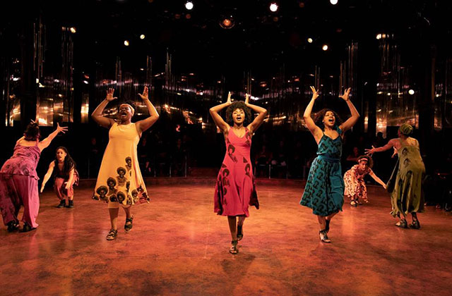"for colored girls." Seven women of color on stage dressed in blue, orange, brown, red, yellow, green and purple.