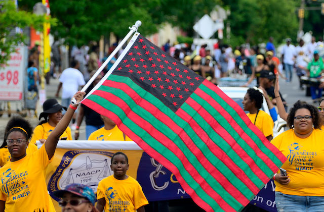 a group of young, Black people wearing yellow shirts march in a parade as they wave a red, black and green flag.