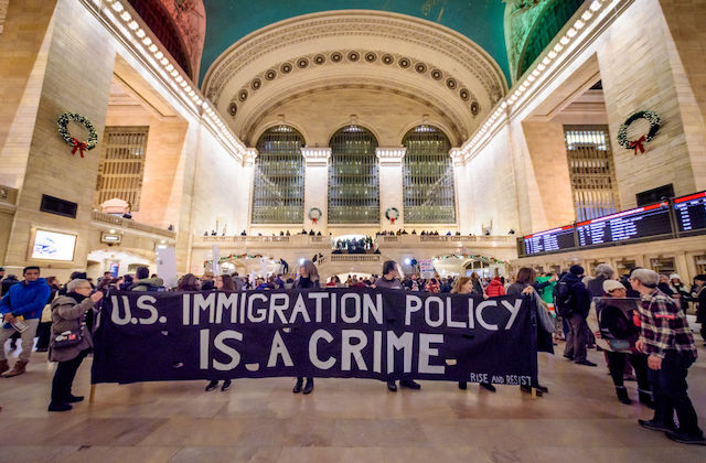 Protestors inside a large train station hold a giant sign that reads, U.S. Immigration Policy is a Crime