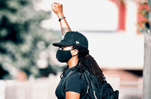 Autostraddle. Black woman wearing black cap, black face mask, black Tee and black back pack with one fist raised in the air.