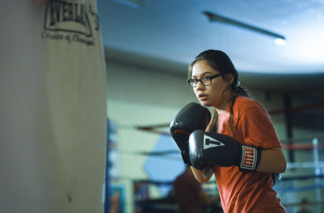 Donna Kipp. Young Blackfeet woman with dark hair, wearing glasses, orange Tee,  black boxing gloves standing in front of a body bag.