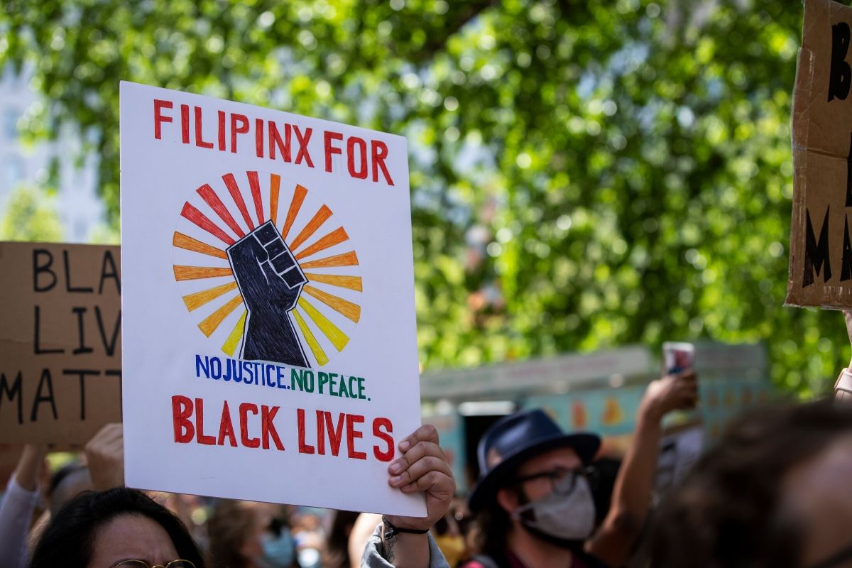 A protester holds a homemade sign that says, "Filipinx For Black Lives No Justice No Peace" with a black power fist and rainbow coming out from the center of the fist