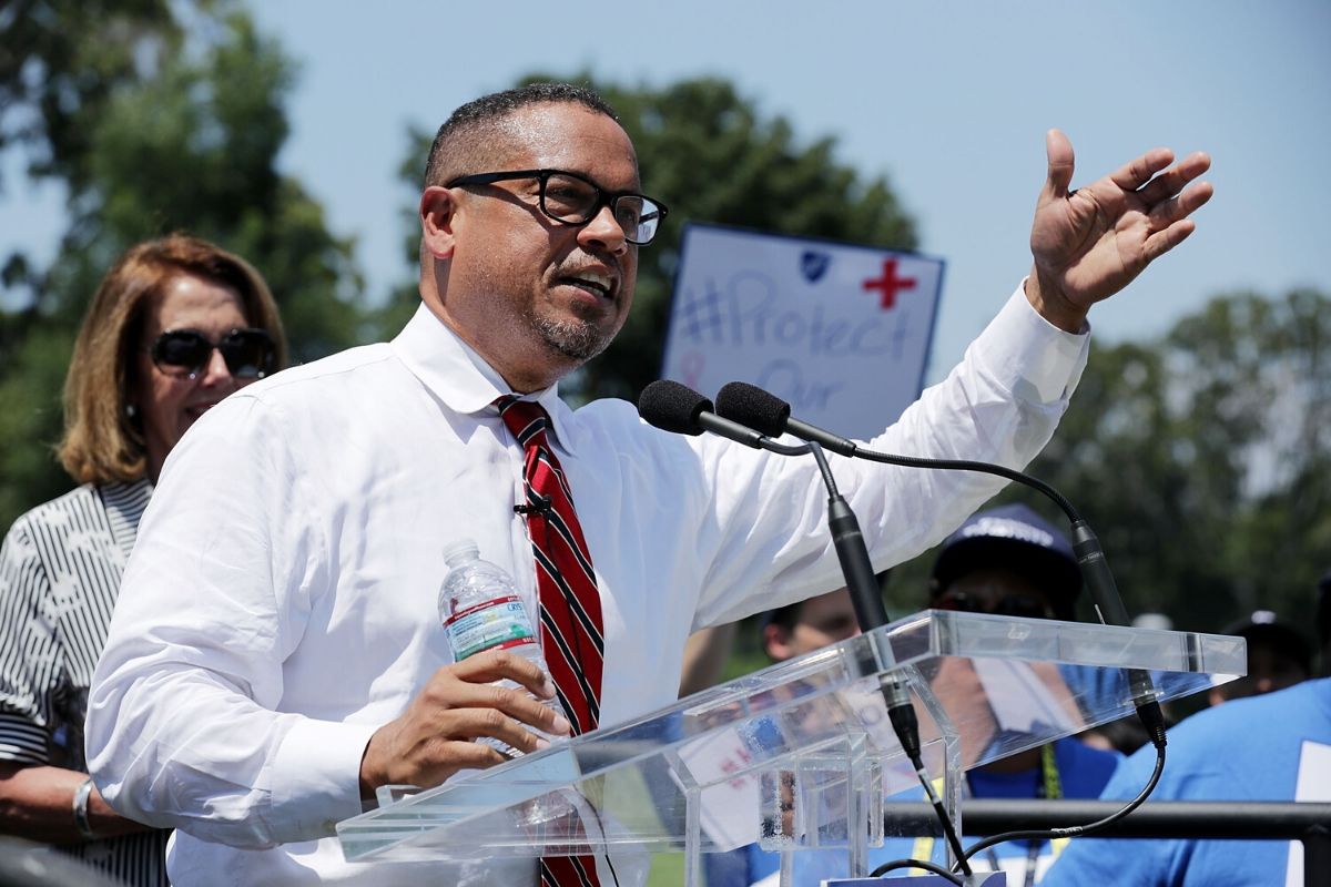 Keith Ellison. African American man with glasses, white shirt and red tie, at podium during the day