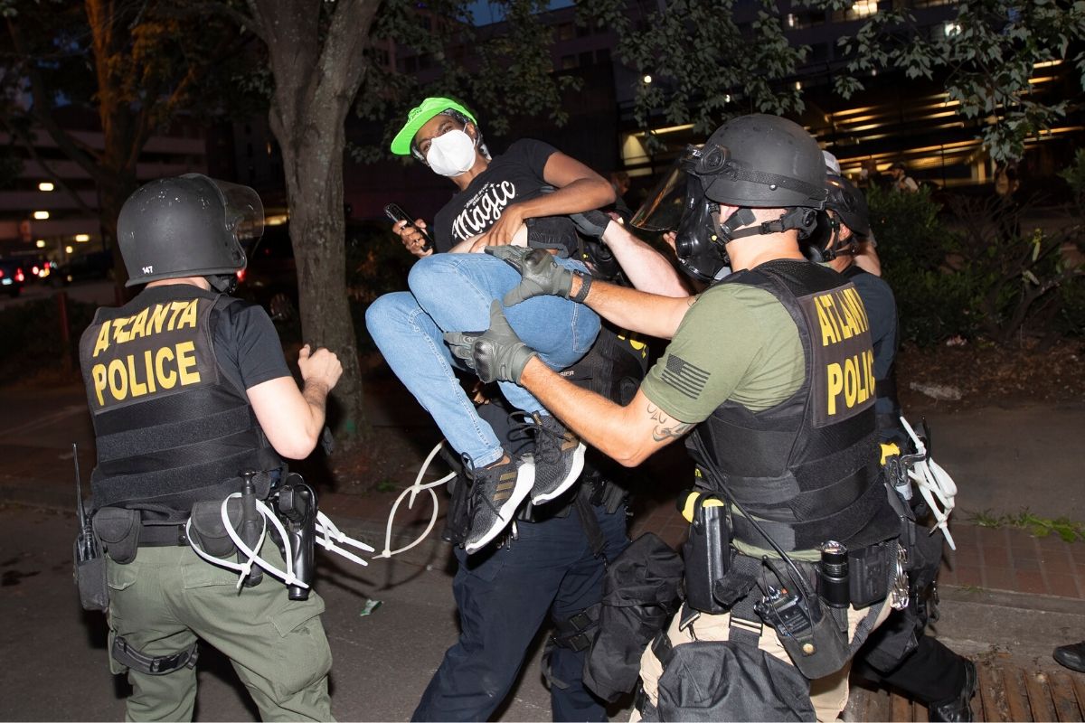 Three military officers in helmets lifting up a Black woman, who has on a neon green cap.