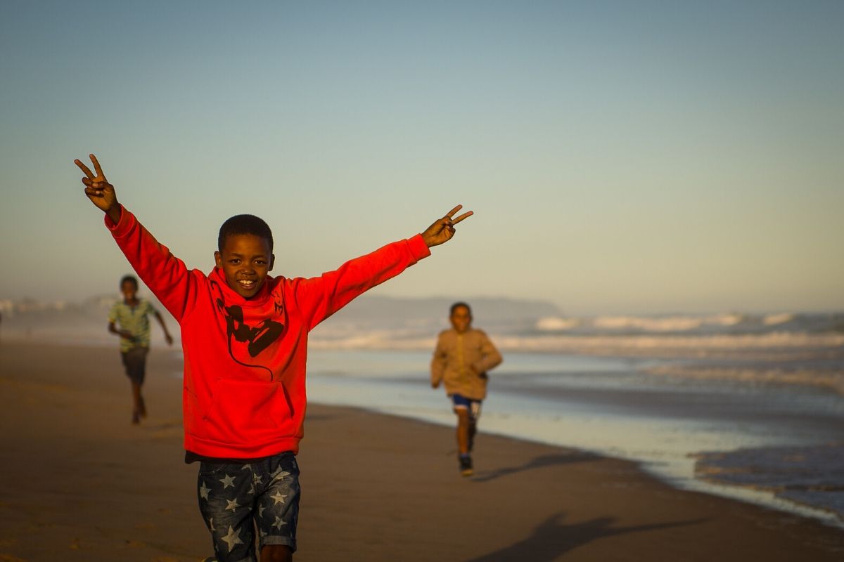 Black Boy Joy. Three black boys running on the beach, the closest, about 9, is smiling from ear to ear.