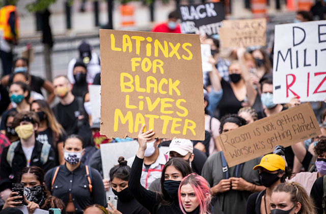 Latinx for BLM. Crowd of protestors where one woman holds up sign with yellow letters reading: "LATiNXS FOR BLACK LiVES MATTER"