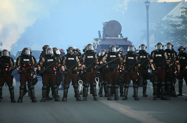 Minneapolis PD. A row of police in black uniforms wearing riot gear, standing in front of an armored vehicle.