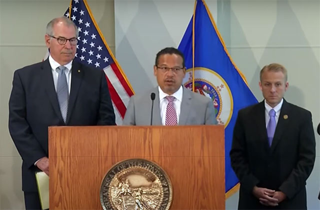 MPD Charges. Black man wearing glasses and gray suit stands in front of podium next to two White men wearing black suits.