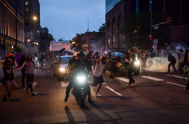 Demonstrators run through a city street as police officers deploy tear gas behind them.