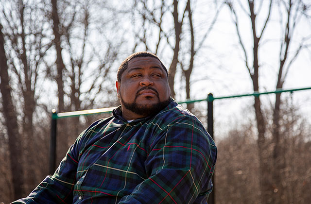 Robert Williams. Black man with short dark hair and facial hair wearing a blue plaid shirt in a park.