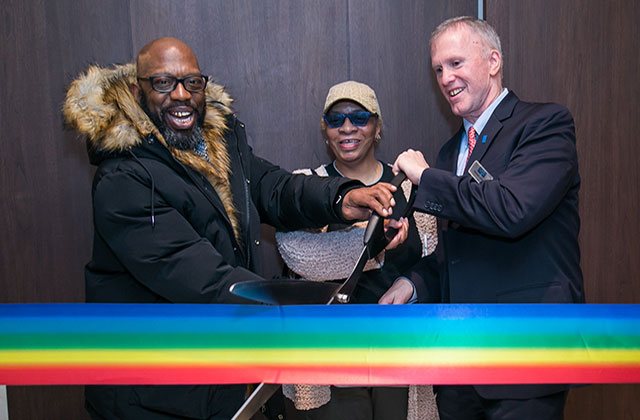 Stonewall House. Ribbon in rainbow colors being cut by Black man with bald head and dark glasses wearing dark winter coat, Black woman wearing tan cap and White man with gray hair wearing dark suit.