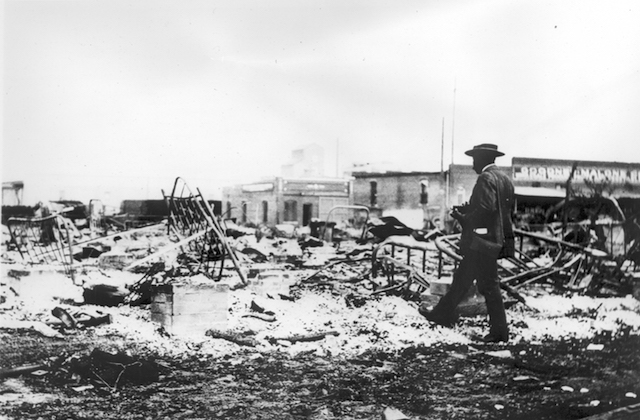 An African-American man with a camera looks at the skeletons of iron beds which rise above the ashes of a burned-out block after the Tulsa Race Massacre, Tulsa, Oklahoma, June 1921