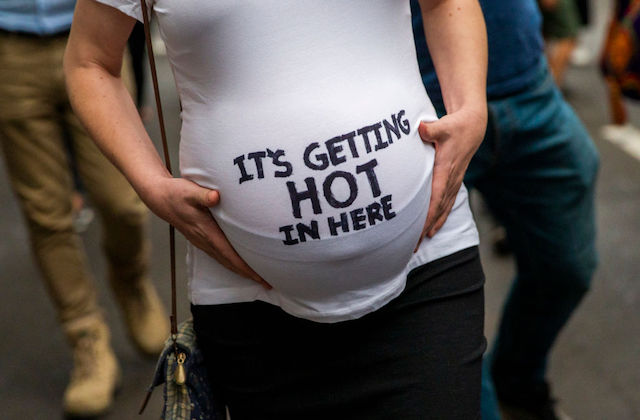 A pregnant stomach is cradled by arms and hands, in a white t-shirt that reads "It's Getting Hot in Here."