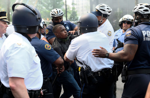 A Black man wearing a black shirt is struggling with at least seven police officers as they surround him.