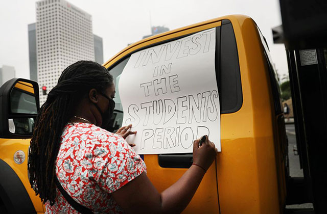 Black-student. Black woman with long braids and red/white dotted shirt writes on sign, "Invest in students period."