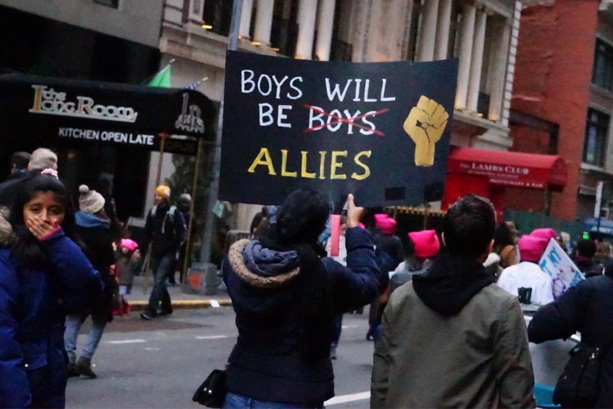 Image of a protest march where a young Brown girl is covering her mouth, and a woman has a sign reading "Boys Will be Boys Allies" with the second "Boys" crossed out