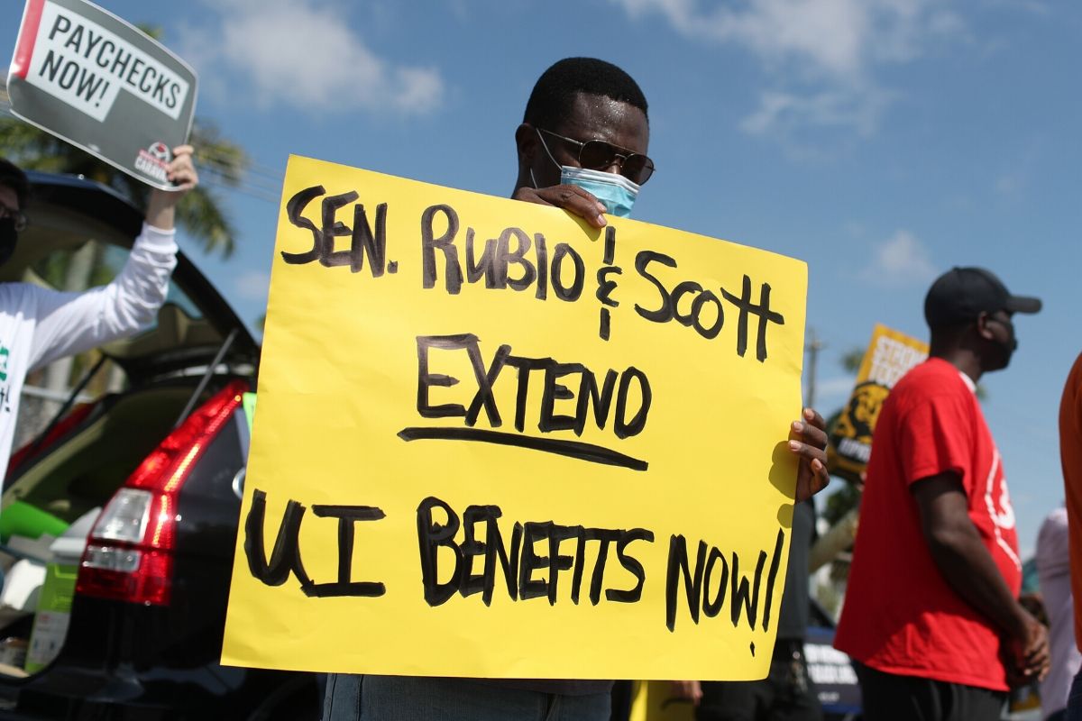 Protest. Black man with mask on holding a yellow sign saying "Sen. Rubio & Scott Extend UI Benefits Now!"