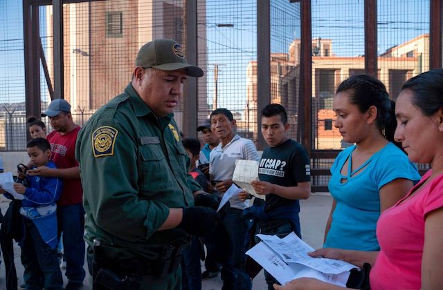 A man wearing a green uniform reviews papers held by two women as a group of men stand in the background.
