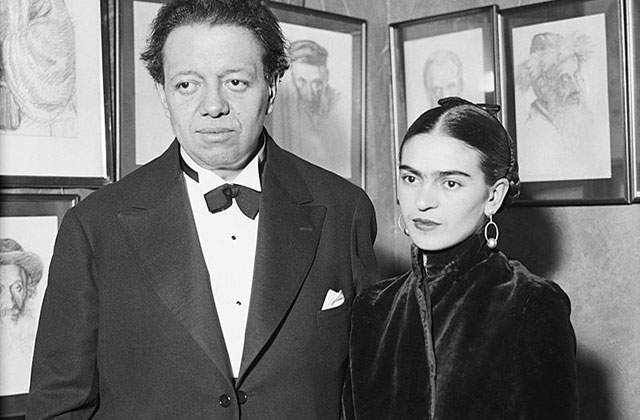 Diego Rivera and Frida Kahlo. Black and white photo of tall Latinx man in black suit with white shirt stands next to smaller Latinx woman wearing black.
