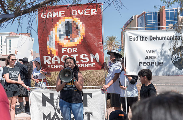 Imagining the Indian. Woman on stage wearing black tee and blue jeans holding a bullhorn in front of a huge banner reading: Game Over for Racism.