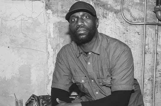 Malik Basit. Black and white photo of Black man wearing a hat and dark shirt.