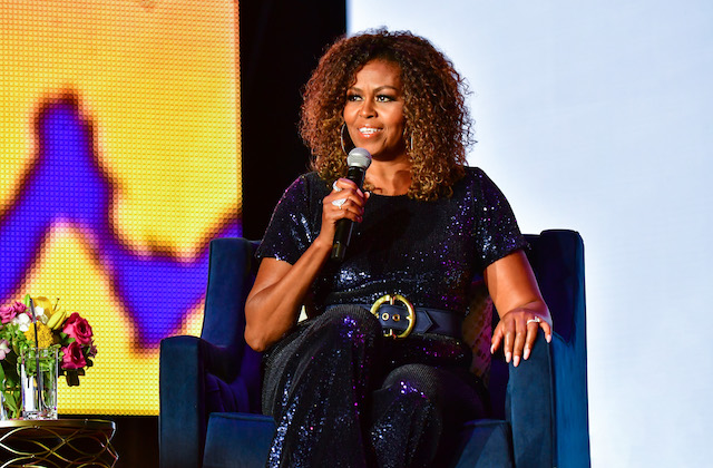 Michelle Obama sits on a blue chair on stage holding a microphone near her face.