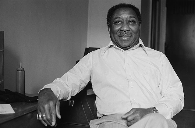 Muddy Waters. Black and white archival photo of Black man with short dark hair wearing white shirt and tan pants.