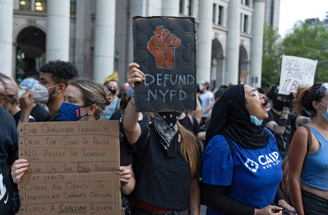 A protester with a scarf covering his face is seen displaying placard reading "Defund NYPD"