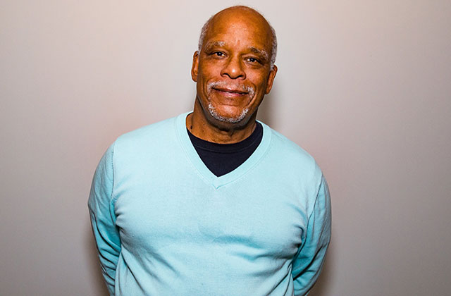 Stanley Nelson. Middle aged Black man with bald head and gray beard wearing a teal colored sweater and black Tee.
