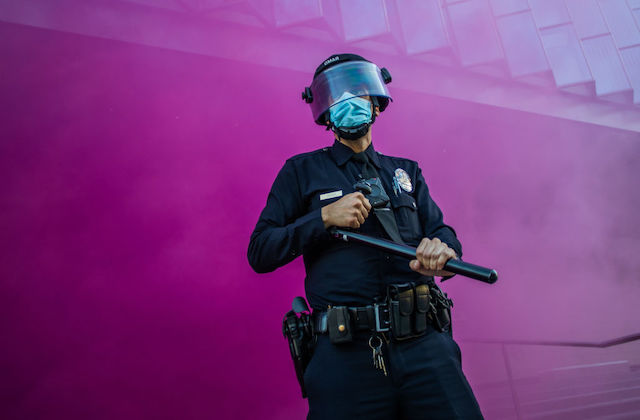 A uniformed police officer stands in front of a purple wall of smoke as they hold a baton and cover their face with a mask and shield.