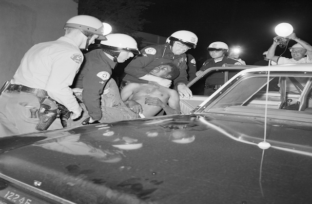 Black and white photograph of police officers forcing a Black man into a police car during second night of the Watts rebellion.